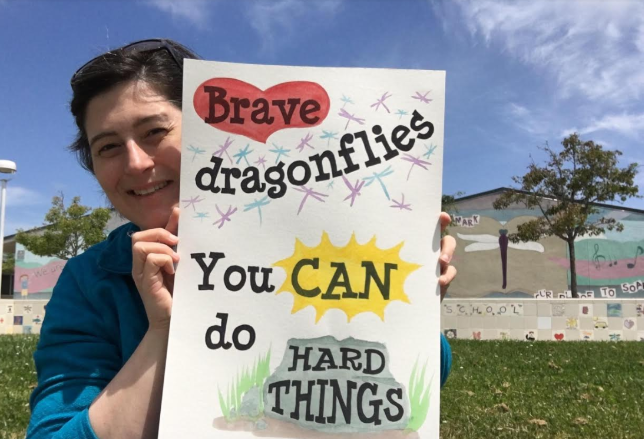 School librarian holding a sign that says "Brave Dragonflies, you can do hard things!"